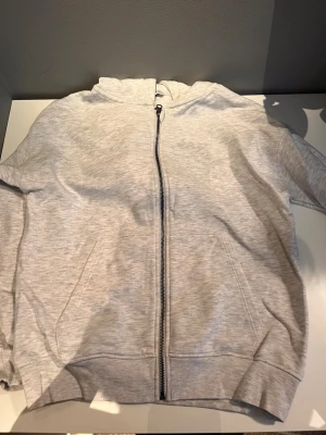 Ljusgrå zip-hoodie med huva - En stilren ljusgrå hoodie med dragkedja framtill och huva. Tröjan har ribbade muddar vid ärmslut och nederkant samt två fickor på sidorna. Perfekt för en avslappnad look och enkel att matcha med andra plagg.