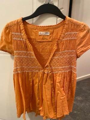 Orange kortärmad blus med brodyr - Söt orange blus med korta ärmar och vita broderade detaljer över bröstet och ryggen. Blusen har en v-ringning med knytband framtill och smock upptill för en avslappnad passform. Tillverkad i ett lätt och luftigt tyg, perfekt för varma dagar.