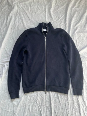 Mörkblå stickad ziptröja Selected Homme - Snygg mörkblå stickad tröja från Selected Homme med hel dragkedja framtill och hög krage. Tröjan har ribbade muddar vid ärmslut och nederkant, perfekt för lager på lager. Materialet är mjukt och ger en clean look.