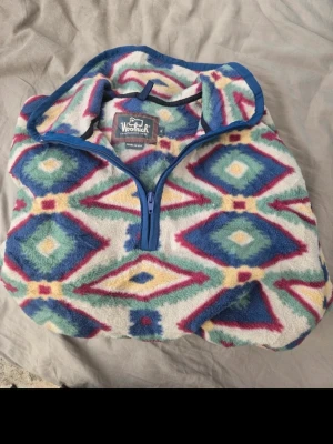 Fleecetröja med mönster från Woolrich - Säljer en färgglad fleecetröja från Woolrich med halv dragkedja och grafiskt diamantmönster i blått, grönt, vitt och vinrött. Tröjan har hög krage och långa ärmar, perfekt för kalla dagar. Materialet är mjuk fleece som håller dig varm.
