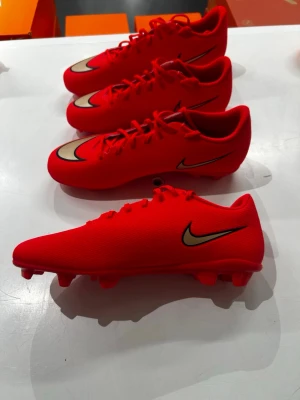 Röda Nike fotbollsskor med guldlogga - Snygga fotbollsskor från Nike i en intensiv röd färg med guldfärgad Swoosh på sidan och ovansidan. Skorna har snörning och är designade för spel på gräs. Tillverkade i ett slitstarkt syntetmaterial som ger bra bollkontroll och passform.