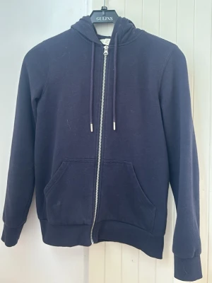 Mörkblå zip hoodie med huva - En enkel och stilren mörkblå hoodie med dragkedja framtill och två stora fickor. Hoodien har justerbar huva med snören och ribbade muddar vid ärmar och nederkant. Perfekt för en avslappnad look.
