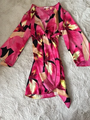 Blommig omlottklänning från H&M - Säljer en färgstark blommig omlottklänning från H&M i rosa, svart, beige och gul. Klänningen har trekvartsärm, knyt i midjan och v-ringad hals. Perfekt för sommarens alla tillfällen och ger en riktigt fräsch vibe. Aldrig använd - tror nypris låg runt 400-500 kr! Perfekt för sommaren/våren och kommer vara perfekt nu i sensommaren.