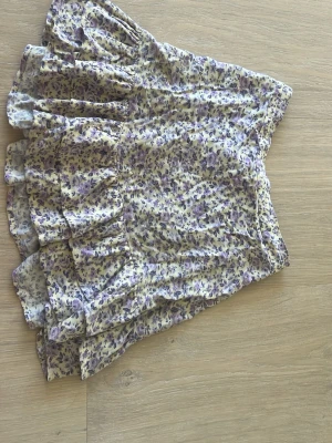 Blommig volangkjol i lila och gult - Söt kortkjol med blommigt mönster i lila och gult. Kjolen har två volanger och resår i midjan där bak för en bekväm passform. Använd ganska mycket men väldigt bra skick. Storleken är 134-140 men passar större.
