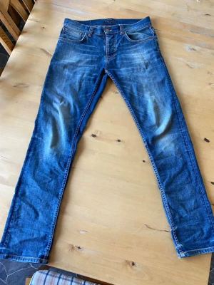 Blå nudie jeans  - Säljer ett par blå nudie slim fit jeans med snygga slitningar och tvättade detaljer. Klassisk femficksmodell med knappgylf och kontrastsömmar. Jeansen har en modern passform och är perfekta för en avslappnad stil. Materialet är denim i bomull. W32 L32 se i sista bilden. Har ni funderingar eller vill ha mer info om jeansen är det bara att höra av sig. Priset är inte hugget i sten