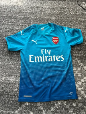 Blå Arsenal matchtröja Puma - Snygg blå Arsenal matchtröja från Puma med korta ärmar och rund hals. Tröjan har Arsenal-logga på bröstet, Puma-loggor på bröst och ärmar samt Fly Emirates-tryck framtill. Tillverkad i lätt och ventilerande material, perfekt för fotbollsträning.
