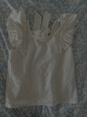 Vit broderad linnetopp med volang - Supersöt vit linnetopp med broderade detaljer och volangprydda axelband. Toppen har en stor rosett i ryggen och är ärmlös, perfekt för sommarens alla tillfällen. Materialet känns luftigt och lätt.