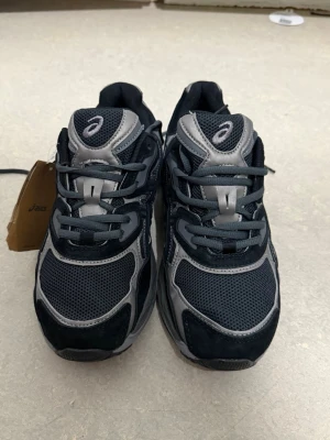 Svarta och grå sneakers från Asics - Snygga sneakers från Asics i svart och grått med mesh- och syntetpaneler. Skorna har en sportig design med tydliga logodetaljer på sidan och tungan. Tjock sula för extra komfort och stabilitet. Perfekta för dig som gillar streetwear och sportig stil.