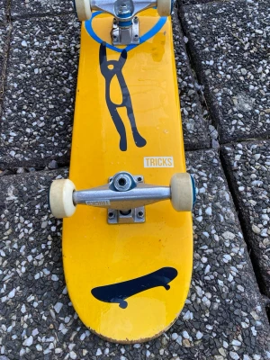 Gul skateboard med svart motiv - Cool skateboard med gul undersida och svart motiv av en figur samt en skateboard. Brädan har svarta grip tape på ovansidan och vita hjul. Märkt med 'TRICKS' på undersidan. Perfekt för dig som gillar att cruisa eller öva tricks i skateparken.
