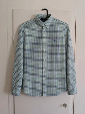 Polo Ralph Lauren rutig skjorta - Långärmad skjorta från Polo Ralph Lauren med små rutor i ljusgrönt, blått och vitt. Mörkblå broderad Polo-logga på bröstet och klassisk button-down-krage. Stilren modell och färgkombination som är lätt att matcha. 📏 Storlek: L (14-16) ✨ Skick: Mycket bra utan defekter! Hör av dig vid frågor eller om du vill ha mått/fler bilder! 📦
