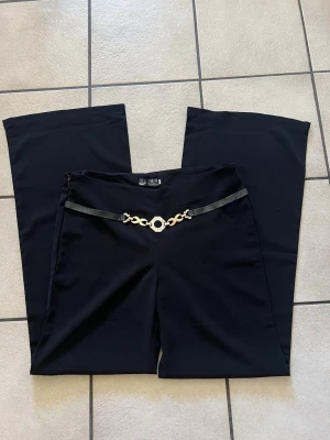 Svarta utsvängda byxor med kedjedetalj - Snygga svarta byxor med utsvängda ben och en dekorativ kedjedetalj i guld och svart band i midjan. Byxorna har hög midja och en stilren look som passar till många olika outfits. Materialet ser ut att vara mjukt och stretchigt för extra komfort.