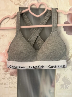 Grå bh från Calvin Klein - Snygg grå bh från Calvin Klein med bred vit resår och svart logga. Bh:n har korsade axelband i ryggen och är tillverkad i mjukt, stretchigt material för extra komfort. Perfekt för en aktiv vardag eller som ett bekvämt basplagg.