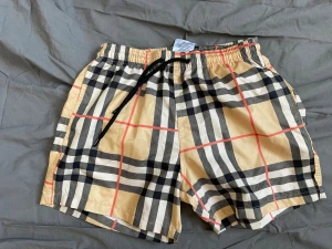 Burberry rutiga badbyxor beige - Snygga badbyxor från Burberry i klassiskt beige rutmönster med svarta, vita och röda detaljer. Elastisk midja med svart dragsko och innerbyxa i mesh för extra komfort. Perfekta för stranden eller poolen.