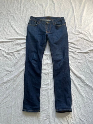Mörkblå jeans från Nudie Jeans - Säljer ett par klassiska mörkblå jeans från Nudie Jeans med raka ben och snygga kontrastsömmar. Jeansen har fem fickor och är tillverkade i slitstark denim. Perfekta för dig som gillar tidlös och enkel stil.