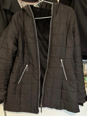 Svart pufferjacka med huva - Snygg svart pufferjacka med huva och två dragkedjeförsedda fickor framtill. Jackan har quiltat mönster och dragkedja hela vägen fram. Perfekt för kyliga dagar och ger en avslappnad streetwear-look.