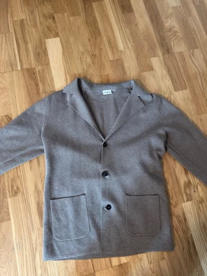 Beige kavaj från John Henric - Stilren beige kavaj från John Henric med två stora framfickor och tre knappar framtill. Kavajen har klassisk krage och är tillverkad i ett mjukt, stickat material som ger en avslappnad men ändå dressad look.