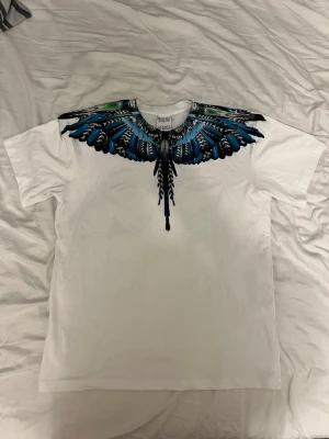 Marcelo Burlon t-shirt - Säljer en vit t-shirt med ett fett blått och svart fjädermönster runt halsen, både fram och bak. T-shirten har normal passform och korta ärmar. Perfekt för dig som vill sticka ut med en unik design.