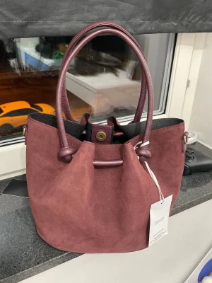 Vinröd bucket bag i mocka - Snygg vinröd bucket bag i mjuk mocka med matchande handtag i läderimitation. Väskan har rundad form, magnetknäppning och detaljer i guld. Perfekt storlek för det viktigaste och ger en trendig touch till din outfit.