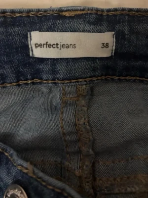 Blå slitna jeans från Gina - Säljer ett par blå jeans från Perfect Jeans i storlek 38. Jeansen har en klassisk femficksmodell, slitningar och hål på ena låret för en trendig look. Materialet är denim med snygg tvätt och kontrastsömmar. Perfekta för dig som gillar en avslappnad stil.