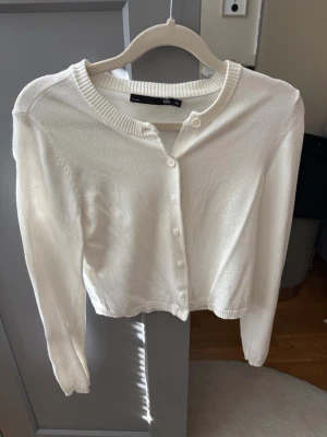 Vit ribbad kofta från 157 - Klassisk vit kofta från 157 med rund halsringning och ribbade detaljer vid krage, ärmslut och nederkant. Koftan har knäppning framtill med små vita knappar och är tillverkad i ett mjukt, tunt material. Perfekt att slänga över en topp eller klänning.