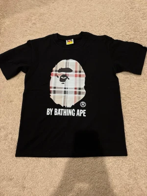 Svart BAPE t-shirt med rutigt tryck - Svart t-shirt med klassiskt BAPE-tryck framtill där apan är i rutigt mönster. Texten 'BY BATHING APE' under motivet. T-shirten har rund hals och korta ärmar, perfekt för en streetwear-look.