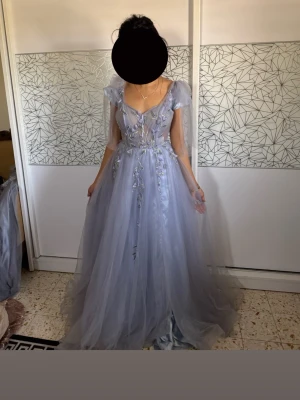 Ljusblå långklänning med blommor - Magisk ljusblå långklänning i tyll med v-ringning och transparenta detaljer. Perfekt för bal eller bröllop osv. Endast använd en gång prislapp kvar. Enda klänning i världen finns inte liknande. Det är lite smutsig längst ner vid fötterna men det kan man klippa bort eller tvätta för hand om man vill, det syns inte så mycket. Genomskinlig runt magen men jag valde ha vit line under. Köph för 6000kr