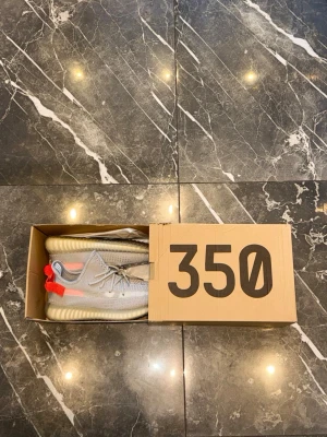 Grå Yeezy 350  - Säljer ett par Yeezy 350 sneakers i ljusgrått med stickad ovandel och tydliga röda detaljer på hälen. Skorna har genomskinlig sula och snörning framtill. Perfekta för dig som vill ha en clean och modern look.