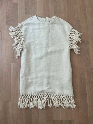 Kortklänning med fransar - En offwhite klänning i bohem stil med fransar. Storlek XS Dragkedja på baksidan. Fickor.  Material: 49% bomull, 44% polyester, 7% viskos. Fransarna är 100% bomull 