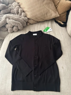 Svart klassisk kofta med knappar - Stilren svart kofta med långa ärmar och knäppning framtill. Koftan har en enkel krage och ribbade muddar vid ärmslut och nederkant. Perfekt att slänga över en t-shirt eller blus för en clean look. Materialet ser ut att vara mjukt och tunt, troligen bomull eller viskos.