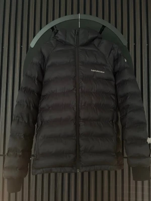 Svart pufferjacka Peak Performance - Säljer en svart pufferjacka från Peak Performance med huva och dragkedja framtill. Jackan har två sidofickor med dragkedja och logga på bröstet. Perfekt för kalla vinterdagar.