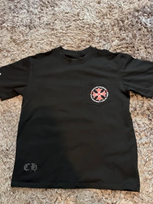 Svart Chrome Hearts t-shirt med tryck - Svart t-shirt från Chrome Hearts med rund hals och kort ärm. På bröstfickan finns ett stort tryck med röd och vit logga och texten 'Chrome Hearts Made in Hollywood USA'. Liten CH-detalj nere vid fållen.