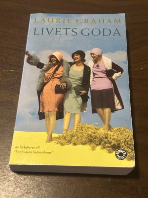 Livets Goda av Laurie Graham - En pocketbok med titeln 'Livets Goda' av Laurie Graham. Omslaget visar tre kvinnor i 1920-talskläder mot en blå himmel. Boken är tryckt i färg med mjukt omslag och tillhör genren skönlitteratur. Perfekt för dig som gillar humor och historiska romaner.