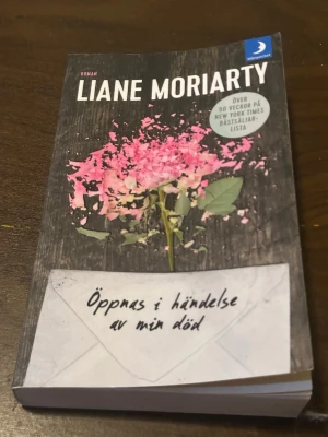 Öppnas i händelse av min död - Romanen 'Öppnas i händelse av min död' av Liane Moriarty är en pocketbok med ett omslag i svart och rosa, prydd med en krossad blomma och ett brev. Boken är utgiven av Månpocket och handlar om hemligheter och familjedrama.