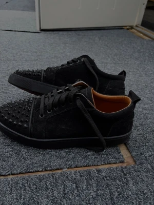 Svarta sneakers med nitar - Säljer ett par svarta sneakers i mocka-look med coola svarta nitar på tån. Skorna har svarta skosnören och en låg siluett. Insidan är ljusbrun och sulan är svart. Perfekta för dig som vill sticka ut med din stil.