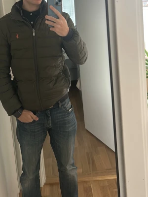 Olivgrön pufferjacka med dragkedja - Den har lite slitningar därför säljs så billigt Säljer en olivgrön pufferjacka med dragkedja framtill och hög krage. Jackan har en liten orange logga på bröstet och quiltad design som ger en schysst streetstil. Perfekt för kalla dagar och riktigt skön att bära.