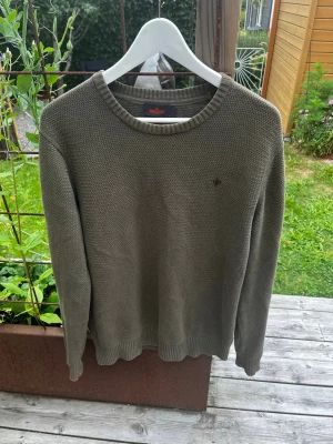 Mörkgrön stickad tröja Morris - Storlek: Medium, Bra skick!Snygg olivgrön stickad tröja med rund hals och diskret broderad detalj på bröstet. Tröjan har en grovstickad struktur och ribbade muddar vid ärmslut och nederkant. Perfekt för dig som gillar en stilren och enkel look.