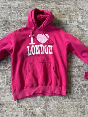 Rosa hoodie med I Love London-tryck - Säljer en rosa hoodie med vit text och ett hjärta på bröstet där det står 'I ♥ LONDON'. Hoodien har huva med dragsko och en stor magficka framtill. Perfekt för dig som gillar London eller vill ha en färgglad och bekväm tröja.
