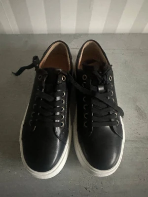 Svarta sneakers med vita sulor - Stilrena svarta sneakers med vita sulor och svarta skosnören. Skorna har metallfärgade öljetter och en låg profil. Insidan är brun och materialet ser ut att vara en mix av läder och textil. Perfekta för en clean och enkel look.