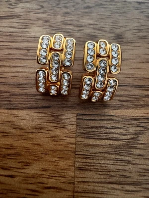 Guldtonade örhängen med strass - Säljer ett par chunky örhängen i guldtonad metall med geometrisk design och flera rader gnistrande klara stenar. Örhängena har push back-lås och en cool, retroinspirerad look som verkligen sticker ut. Perfekta för dig som gillar statement-smycken.