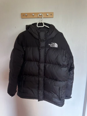 Svart dunjacka från The North Face - Säljer en svart dunjacka från The North Face med huva och logga på bröstet och ryggen. Jackan har en puffig design med breda kanaler och dragkedja framtill. Perfekt för kalla vinterdagar och riktigt varm tack vare dunfyllningen.