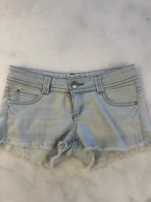Ljusblå jeansshorts med fransig kant - Säljer ett par ljusblå jeansshorts med fransig kant och klassisk femficksdesign. Shortsen har bälteshällor, knapp och dragkedja framtill samt råa kanter nedtill för en avslappnad look. Perfekta för varma dagar!