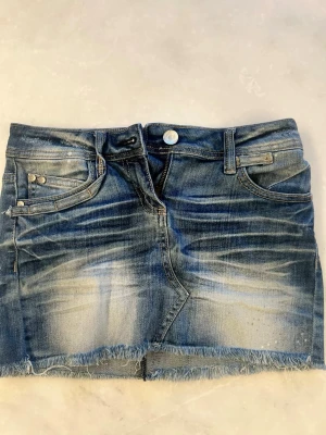 Blå jeanskjol med fransig kant - Säljer en blå jeanskjol i klassisk denim med slitningar och fransig nederkant. Kjolen har fem fickor, bälteshällor och knappstängning framtill. Snygg tvättad look och rå kant nedtill för en trendig vibe.