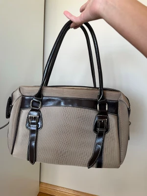 Beige handväska med bruna detaljer - Stilren beige handväska med mörkbruna detaljer och dubbla handtag. Väskan har dekorativa spännen framtill och dragkedja upptill. Insidan är rymlig med innerfack för småsaker. Materialet är en mix av tyg och konstläder vilket ger en snygg kontrast.