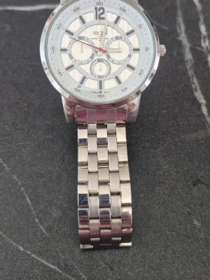 Silverfärgad klocka från Regal - Stilren klocka från Regal med silverfärgat metallarmband och vit urtavla. Klockan har flera dekorativa subdials och tydliga siffror. Perfekt accessoar för dig som gillar en klassisk look med modern touch.