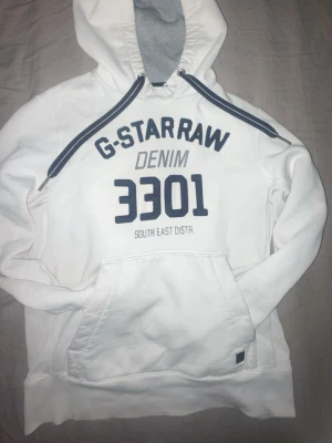 Vit hoodie från G-Star RAW - Vit hoodie från G-Star RAW med stor blå text 'G-STAR RAW DENIM 3301' på bröstet. Hoodien har en stor känguruficka, dragsko i huvan och mjukt, fleecefodrat innerfoder. Perfekt för en avslappnad streetwear-look.