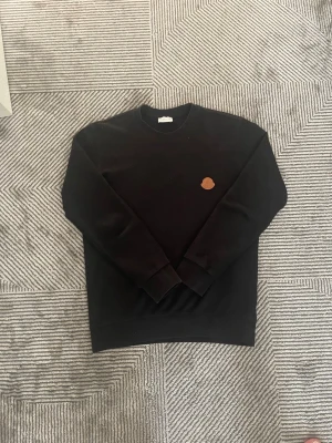 Svart sweatshirt från Moncler - Stilren svart sweatshirt från Moncler med rund halsringning och ett brunt Moncler-märke broderat på bröstet. Tröjan har långa ärmar och ribbade muddar vid ärmslut och nederkant. Perfekt för en clean och avslappnad look.
