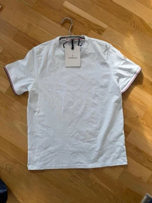 Vit Moncler t-shirt med logga - Säljer en stilren vit t-shirt från Moncler med klassisk logga på ärmen. T-shirten har korta ärmar och tunna rödblå ränder vid ärmslut och halsringning. Tillverkad i mjuk bomull som känns skön mot huden. Perfekt för en clean och fräsch look.