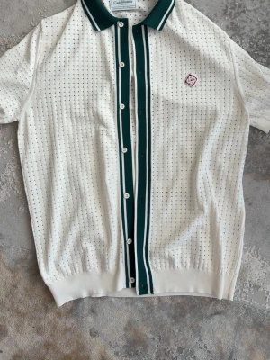 Vit pikéskjorta från Casablanca - Vit pikéskjorta från Casablanca Tennis Club med gröna och vita ränder längs knappraden och kragen. Skit snygg tröja.Små i storlek,  Sitter som XL 