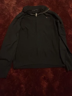 Svart stickad tröja Tommy Hilfiger - Snygg svart stickad tröja från Tommy Hilfiger med halv dragkedja och hög krage. Tröjan har diskret logga på bröstet och ribbade muddar vid hals och ärmslut. Perfekt för lager på lager och enkel att matcha med olika outfits. Står L på lappen men passar mig som är 184 och har S i kläder 