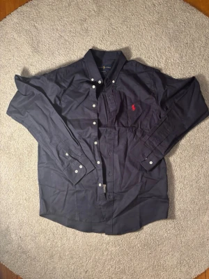Mörkblå skjorta från Ralph Lauren - Klassisk mörkblå skjorta från Ralph Lauren med röd broderad logga på bröstet. Skjortan har lång ärm, knappar framtill och button-down krage. Perfekt för en stilren look och tillverkad i mjuk bomull.